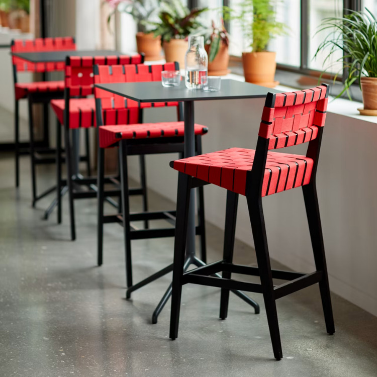 Risom Barstool with a black hardwood frame and vibrant red woven seat, positioned at a sleek table in a bright room.