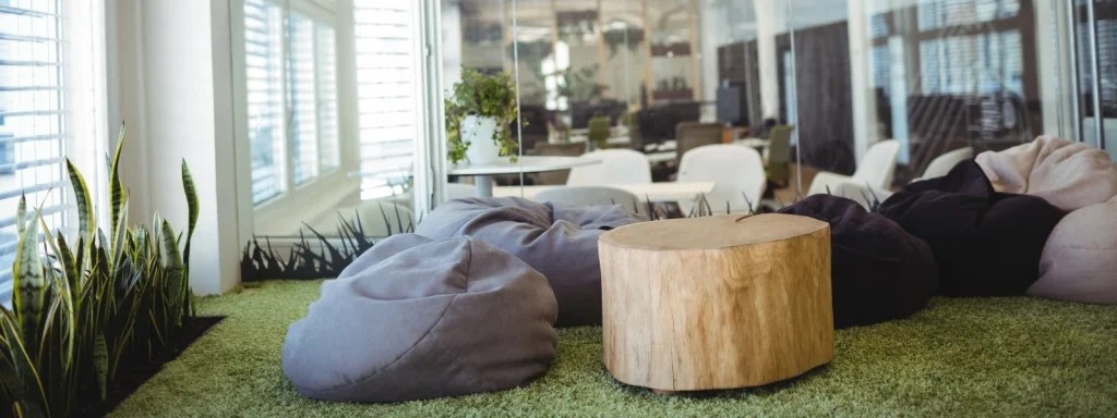 Modern biophilic office lounge with bean bag chairs, a natural wood stump table, and snake plants on green carpet.